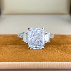 May include: A silver-toned ring featuring a large, rectangular radiant-cut center stone flanked by two smaller trapezoid-cut side stones. The band is thin and encrusted with small, round, clear stones along the visible shank, set in metal.