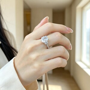 May include: A silver-toned ring featuring a large, rectangular cushion-cut clear center stone flanked by two smaller trapezoidal side stones, set on a thin band encrusted with small, sparkling clear stones along the visible upper shank.