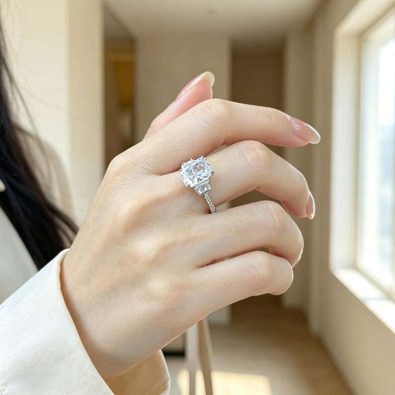 May include: A silver-toned ring featuring a large, rectangular cushion-cut clear center stone flanked by two smaller trapezoidal side stones, set on a thin band encrusted with small, sparkling clear stones along the visible upper shank.
