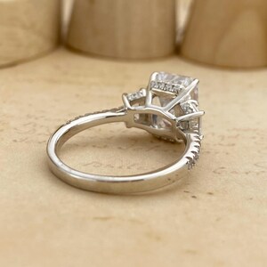 May include: A silver-toned ring featuring a large rectangular center stone held by a prong setting, with smaller round stones embedded along the band and the gallery. The metal band has a polished finish and a smooth, rounded interior profile.