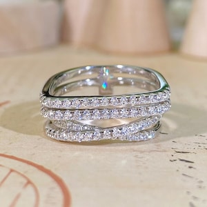 May include: A silver-toned multi-band ring featuring a crossover design. The ring is composed of three distinct bands that intersect, each encrusted with small, clear, sparkling stones along the entire visible surface of the metal bands.