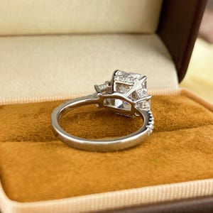May include: A silver-toned ring featuring a large, rectangular-cut center stone flanked by smaller stones on each side. The band is thin and includes a row of tiny, embedded accent stones along the upper shank, showcasing a polished metal finish.
