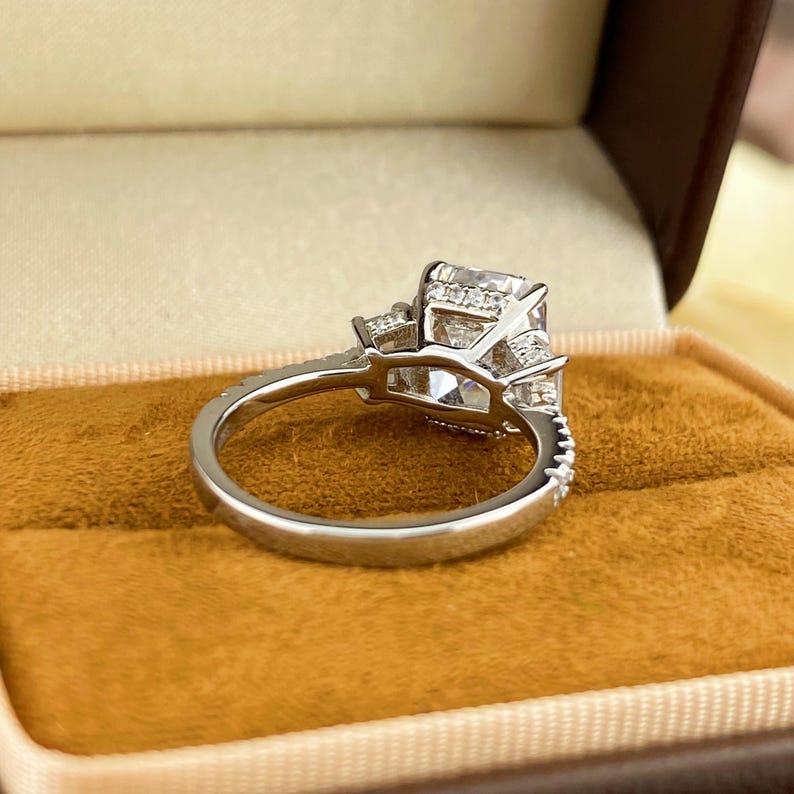 May include: A silver-toned ring featuring a large, rectangular-cut center stone flanked by smaller stones on each side. The band is thin and includes a row of tiny, embedded accent stones along the upper shank, showcasing a polished metal finish.