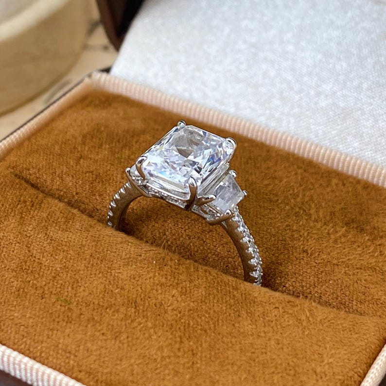 May include: A silver-toned ring featuring a large rectangular radiant-cut center stone flanked by two smaller trapezoid-cut side stones. The band is thin and encrusted with small round pav&eacute;-set stones along the visible outer surface.