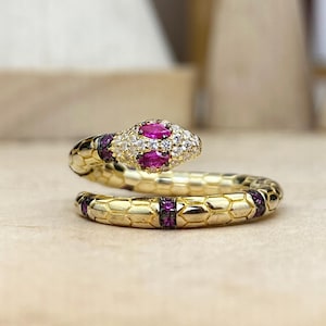 May include: A gold-toned snake ring featuring a textured scale pattern along the band. The snake head is embellished with two marquise-cut pink stones and numerous small clear white stones. Additional small pink stones accent the ring band.