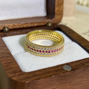 Può includere: Un anello a fascia color oro con tre file parallele di pietre. Le file esterne sono composte da pietre chiare e scintillanti, mentre la fila centrale presenta pietre rosse vivaci. I bordi dell'anello hanno una finitura metallica rigata.