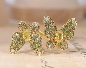 May include: A pair of gold butterfly earrings featuring a central oval yellow gemstone surrounded by small green and clear stones. The wings are detailed with a pavé setting of green and white crystals, creating a vibrant and textured appearance.