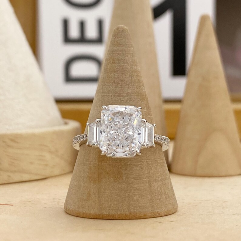 May include: A silver-toned ring featuring a large rectangular radiant-cut center stone flanked by two smaller trapezoid-cut side stones. The thin band is encrusted with small pav&eacute;-set stones along the sides, creating a brilliant, reflective appearance.