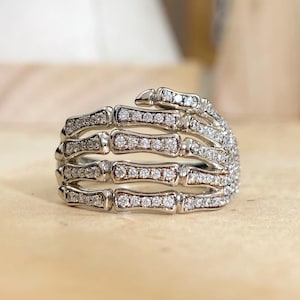 May include: A silver-toned ring shaped like a skeletal hand wrapping around the finger. The metal structure is encrusted with numerous small, clear, sparkling stones that cover the entire surface of the finger bones and knuckles.