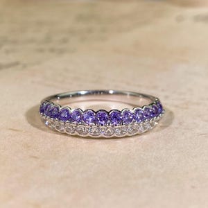 May include: A silver ring with two rows of gemstones. The top row features small, round purple stones, while the bottom row has clear, white stones. The ring is set against a neutral background.