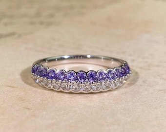 May include: A silver ring with two rows of gemstones. The top row features small, round purple stones, while the bottom row has clear, white stones. The ring is set against a neutral background.