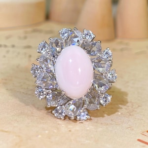 Op de afbeelding: Een zilverkleurige ring met een grote, gladde, ovaalvormige lichtroze cabochonsteen in het midden. De centrale steen is omringd door een halo van meerdere heldere, gefacetteerde, peervormige en ronde stenen in een bloemmotief.