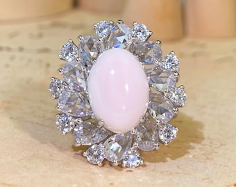 May include: A silver-toned ring featuring a large, smooth, oval-shaped pale pink cabochon stone at the center. The central stone is surrounded by a halo of multiple clear, faceted, pear-shaped and round-cut stones arranged in a floral pattern.