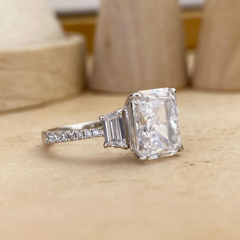 May include: A silver-toned ring featuring a large rectangular radiant-cut center stone flanked by two smaller baguette-cut stones. The band is adorned with a row of small round pav&eacute;-set stones extending along the sides of the metal shank.