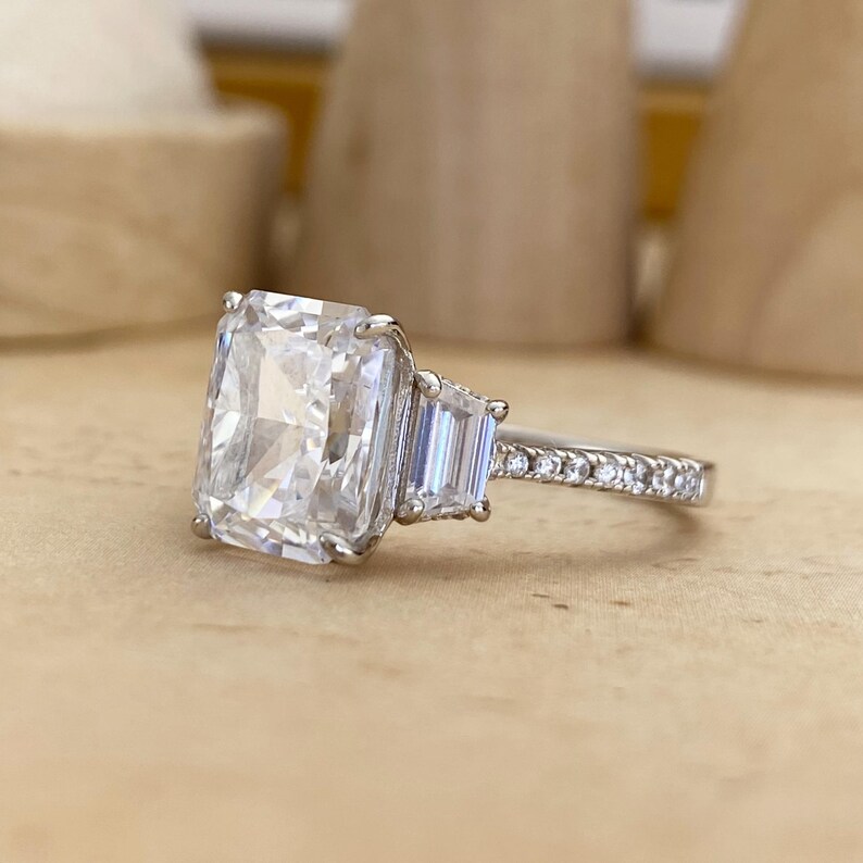 May include: A silver-toned ring featuring a large rectangular radiant-cut center stone flanked by two smaller baguette-cut stones. The band is accented with a row of small round pav&eacute;-set stones extending along the sides of the metal shank.