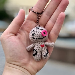 May include: A handmade crochet doll keychain in beige with black stitched details. The doll has a pink button eye and a red heart. The keychain is attached to a bronze-colored ring.