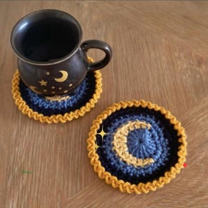 May include: A dark brown mug with a crescent moon and star design sits on a blue and gold crocheted coaster. A second coaster with a crescent moon design is next to the mug. The coasters have a yellow border.