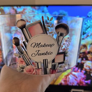May include: A clear, rectangular container with a decorative design featuring makeup brushes, blush, lipstick, and nail polish. The words "Makeup Junkie" are in a circular design. The container is held in a hand.