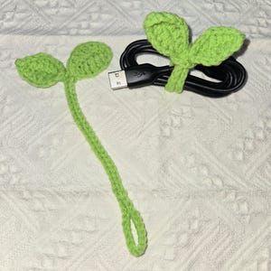 May include: Two green crocheted plant-shaped cable organizers. One is a single stem with leaves at the top, the other is a pair of leaves that can be used to wrap around a cable.