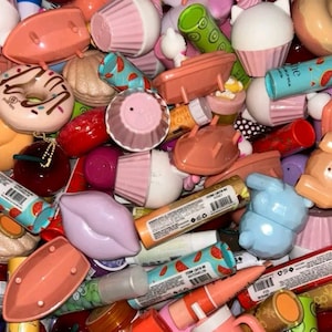 May include: A collection of colorful lip balms and cosmetic containers in various shapes, including cupcakes, boats, and donuts. The assortment features shades of pink, orange, and blue, with some items displaying watermelon and other fruit designs.