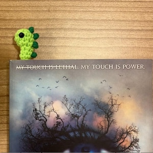 May include: A green crocheted dinosaur bookmark with a black eye and green spikes is placed on top of a book. The book cover has a dark, cloudy sky with bare tree branches and the text "MY TOUCH IS LETHAL. MY TOUCH IS POWER."