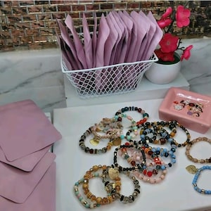 May include: A collection of beaded bracelets in various colors and designs, including gold, blue, and pink. A white wire basket holds pink pouches, and a small pink dish with a decorative design is also present. A small potted plant with pink flowers adds a touch of color.