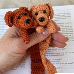 May include: Two handmade crochet dog bookmarks in shades of brown and orange. Each dog has black eyes and a black nose. The bookmarks are held up against a book, showcasing their length and detail. A charming and unique gift for book lovers.