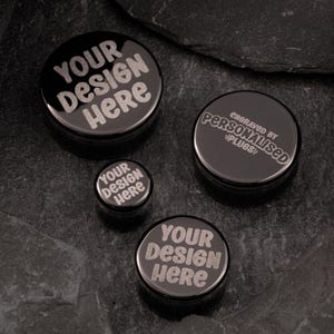 Puede incluir: Cuatro expansores de oreja negros de diferentes tamaños. El expansor más grande tiene el texto "YOUR DESIGN HERE" grabado en él. El segundo expansor más grande tiene el texto "ENGRAVED BY PERSONALISED PLUGS!" grabado en él. Los dos expansores más pequeños tienen el texto "YOUR DESIGN HERE" grabado en ellos.