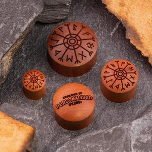 May include: Three wooden ear gauges with a circular design featuring a runic symbol. The gauges are engraved with the text "ENGRAVED BY PERSONALISED PLUGS".