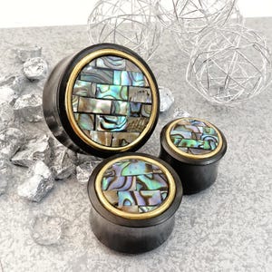 May include: Three wooden ear gauges with a gold trim and a mosaic of iridescent abalone shell inlay. The largest gauge is 3.8 cm in diameter, the middle gauge is 2.5 cm in diameter, and the smallest gauge is 1.9 cm in diameter.