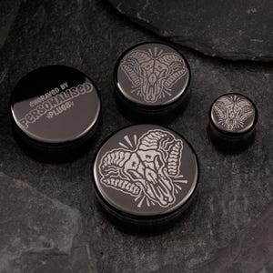 Pode incluir: Conjunto de três expansores de orelha pretos com desenhos de caveira de carneiro gravados. O expansor maior é gravado com o texto "Engraved by Personalised Plugs!"