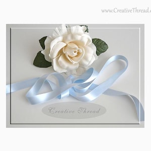 May include: A white silk rose with green leaves and a light blue satin ribbon on a white background. The ribbon is tied in a bow and the text "Creative Thread" is printed on the background.