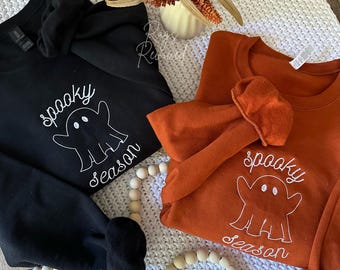 May include: Black and orange sweatshirts with white embroidered "spooky season" ghost design. The sweatshirts are folded on a white knitted blanket, with a decorative fall floral arrangement and wooden beads.