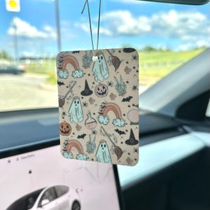 May include: A pink and white car air freshener with a Halloween theme. The design features ghosts, pumpkins, rainbows, and other spooky elements.