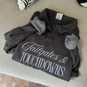 Puede incluir: Camisa de manga larga gris oscuro con el texto bordado "Tailgates & TOUCHDOWNS" en escritura blanca. La camisa tiene cuello y puños grises. La camisa está doblada sobre una superficie gris claro.