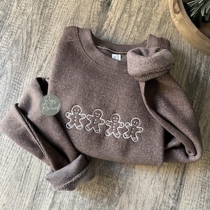 Może przedstawiać: Brązowa bluza z okrągłym dekoltem i długimi rękawami. Bluza posiada haftowany wzór czterech białych ludzików z piernika. Rękawy są podwinięte. Marka "Rags Revived" znajduje się po lewej stronie.