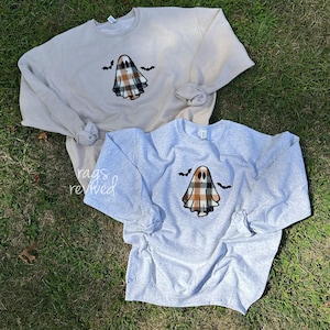 Pode incluir: Duas camisolas de gola redonda, uma bege e outra cinzenta, são exibidas sobre relva verde. Cada camisola apresenta um gráfico de fantasma xadrez com duas silhuetas de morcegos pretos. As camisolas de manga comprida são adequadas para o outono ou Halloween.