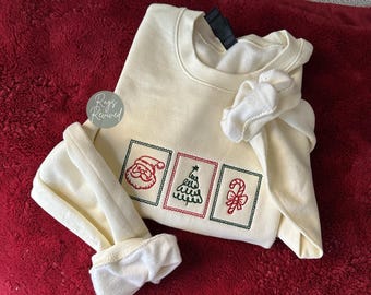 Könnte beinhalten: Cremefarbenes Sweatshirt mit gestickten Weihnachtsmotiven. Das Design umfasst den Weihnachtsmann, einen Weihnachtsbaum und eine Zuckerstange. Das Sweatshirt liegt auf einer roten Oberfläche.
