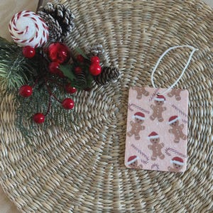 May include: A rectangular air freshener with a light pink background featuring a pattern of gingerbread men wearing Santa hats and candy canes. A white string is attached for hanging. Festive holiday decor.