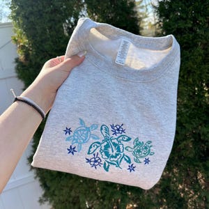 Op de afbeelding: Grijs sweatshirt met een geborduurd ontwerp van drie zeeschildpadden met bloemen en sterren. De schildpadden zijn in tinten blauw en groen.