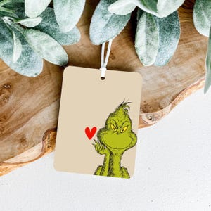 May include: A rectangular tag with a string, featuring a cartoon Grinch character in green with yellow eyes and a red heart. The tag has a beige background and is likely for a gift or decoration.