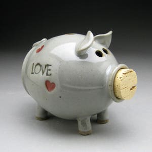 Puede incluir: Una alcancía de cerámica gris con un tapón de corcho para la ranura de monedas. La alcancía tiene la palabra "LOVE" y tres corazones rojos en el lateral.