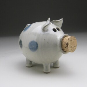 K&ouml;nnte beinhalten: Ein wei&szlig;es Keramik-Sparschwein mit blauen Punkten und einem Korkstopfen f&uuml;r den M&uuml;nzeinschub.