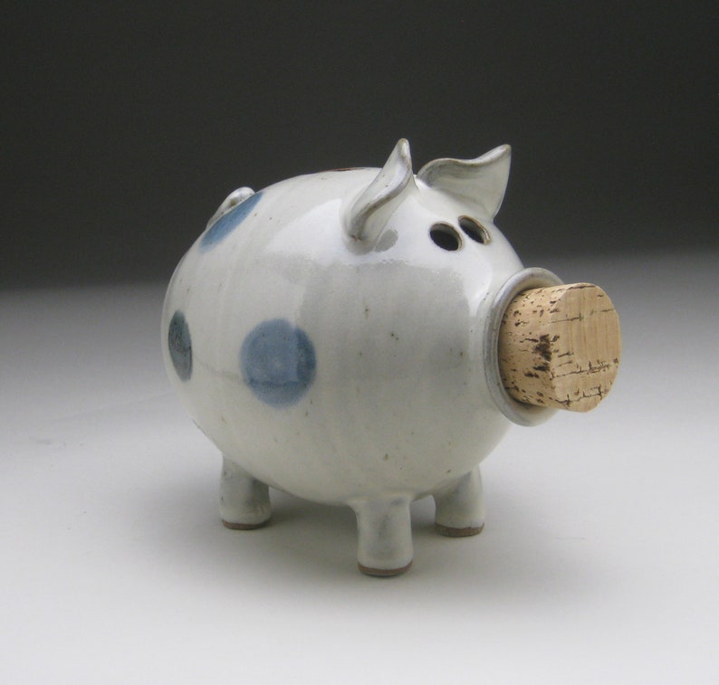 K&ouml;nnte beinhalten: Ein wei&szlig;es Keramik-Sparschwein mit blauen Punkten und einem Korkstopfen f&uuml;r den M&uuml;nzeinschub.