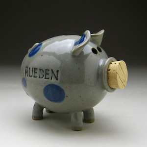 Può includere: Un salvadanaio in ceramica grigia con pois blu e il nome "RUEBEN" inciso. Presenta un tappo di sughero nel muso e quattro zampe corte. Le orecchie sono blu. Un oggetto decorativo per risparmiare.