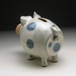 K&ouml;nnte beinhalten: Ein wei&szlig;es Keramik-Sparschwein mit blauen Punkten und einem Korkstopfen. Das Sparschwein hat vier Beine und einen Schwanz.
