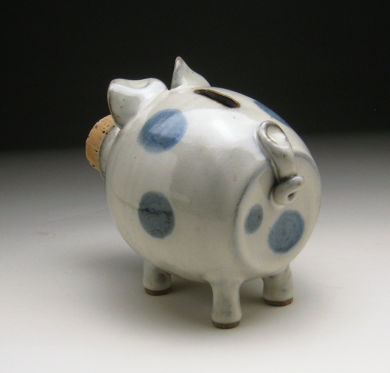 K&ouml;nnte beinhalten: Ein wei&szlig;es Keramik-Sparschwein mit blauen Punkten und einem Korkstopfen. Das Sparschwein hat vier Beine und einen Schwanz.