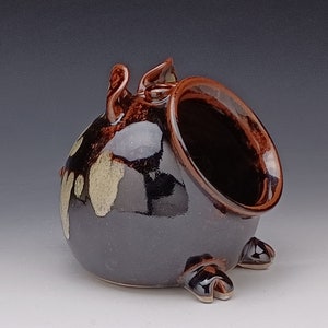 May include: A brown ceramic salt pig with a glazed finish and white speckles. The pig has a large opening on the side for pouring salt.