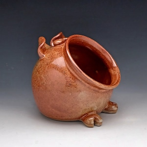 May include: A handmade, ceramic salt pig in a warm, speckled reddish-brown hue. The open-mouthed container features a rounded body, a handle, and three small feet. The interior is a darker shade of brown.