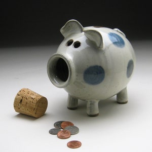 K&ouml;nnte beinhalten: Ein wei&szlig;es Keramik-Sparschwein mit blauen Punkten und einem Korkstopfen. Das Sparschwein hat vier Beine und eine Schnauze. Vor dem Sparschwein liegen mehrere M&uuml;nzen auf dem Tisch.